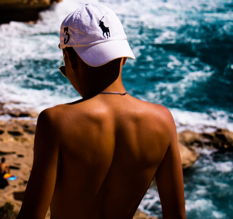 shirtless man with back turned to camera wearing a white polo ralph lauren hat looking at the beach