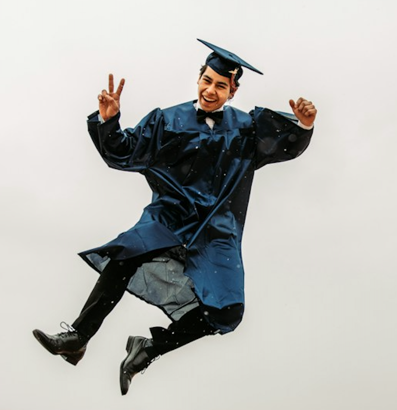 young man in cap, gown, and graduation attire jumping outside