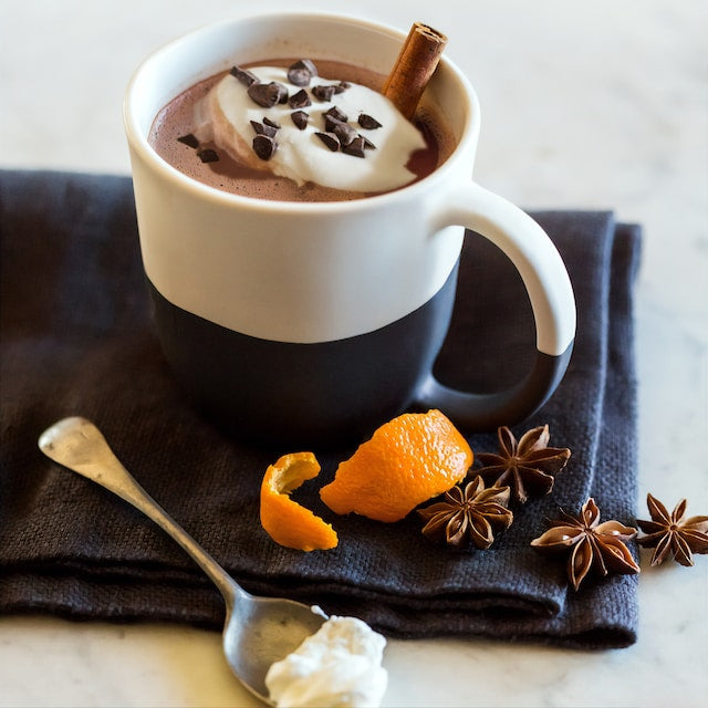mug of hot chocolate with chocolate chips, whipped cream and cinnamon next to orange citrus peel