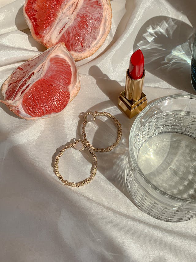 photo of 2 slices of grapefruit next to hoop earrings, red lipstick, and a clear glass